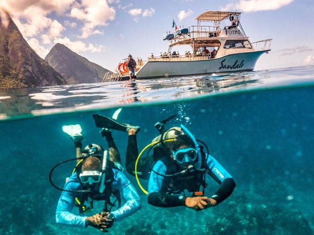 Two scuba divers swim underwater near a dive boat named "Sandals" in clear tropical waters